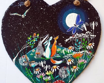 Könnte beinhalten: Eine herzförmige Schiefertafel mit einem fantasievollen Gemälde eines Dachses, Kaninchens und Fuchses, die zum Vollmond aufblicken. Die Szene spielt auf einem Feld voller Blumen mit einer Libelle, die darüber fliegt. Der Hintergrund ist ein sternenübersäter Nachthimmel.