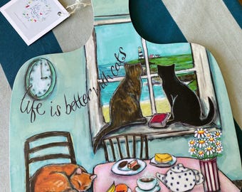Könnte beinhalten: Ein dekoratives Holzschneidebrett mit einer bemalten Szene von drei Katzen in einem Raum mit Fensterblick auf einen Leuchtturm. Der Text "life is better with cats" steht links. Ein Tisch mit Teekanne, Obst und Blumen ist abgebildet.