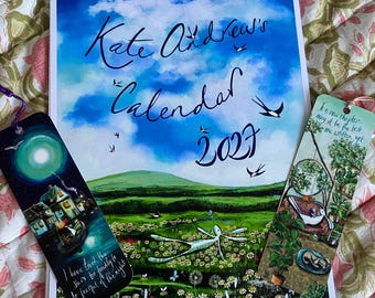 Könnte beinhalten: Ein spiralgebundener Kalender für 2027 mit dem Titel Kate Andrew's Calendar 2027, der einen blauen Himmel mit Schwalben über einer grünen Wiese zeigt, flankiert von zwei Lesezeichen. Eines zeigt eine nächtliche Szene, das andere einen Pflanzenraum.