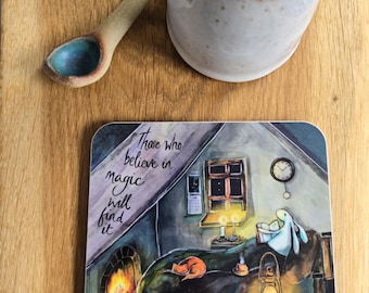 Könnte beinhalten: Ein quadratischer Untersetzer mit einer Illustration eines lesenden Hasen in einem gemütlichen Raum mit Kamin, Laterne und dem Text "Those who believe in magic will find it", dazu eine weiße Keramiktasse und ein kleiner Keramiklöffel.