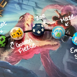 May include: A close-up of a black string with five wooden beads. Each bead is painted with a different design, including a trident, a golden fleece, a blue star, a maze, and an empire state building. The beads are labeled with their corresponding names.