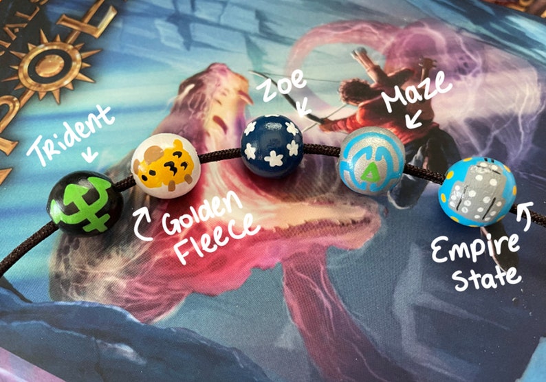 May include: A close-up of a black string with five wooden beads. Each bead is painted with a different design, including a trident, a golden fleece, a blue star, a maze, and an empire state building. The beads are labeled with their corresponding names.