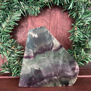 May include: A polished, triangular fluorite crystal with shades of green, purple, and white. The crystal is set against a backdrop of a green wreath and a dark brown surface. The fluorite has a translucent quality.