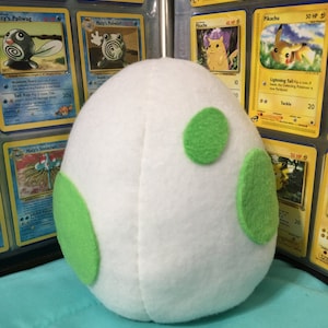May include: A white plush egg with three green felt spots. The egg is sitting on a table with a collection of Pokemon trading cards in the background.