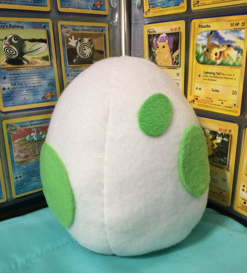 May include: A white plush egg with three green felt spots. The egg is sitting on a table with a collection of Pokemon trading cards in the background.