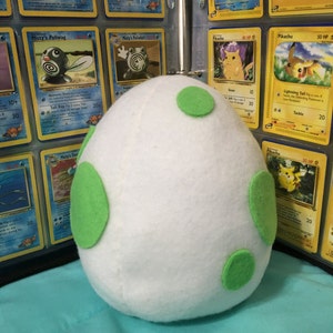 May include: A white plush egg with green felt spots. The egg is sitting on a blue surface in front of a binder of Pokemon trading cards.