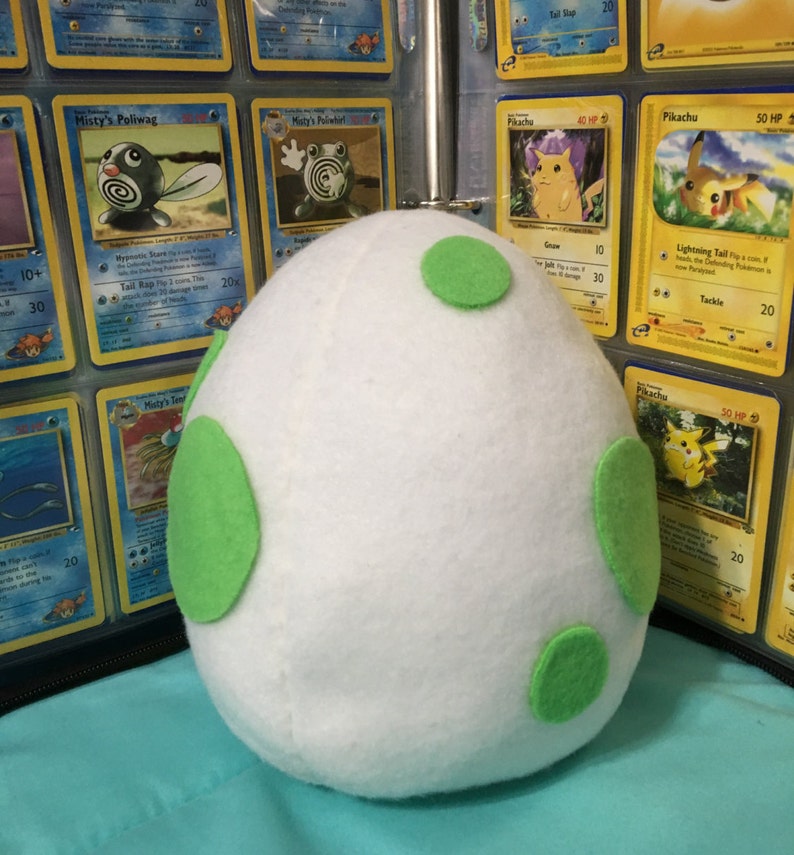 May include: A white plush egg with green felt spots. The egg is sitting on a blue surface in front of a binder of Pokemon trading cards.