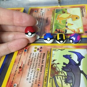 Op de afbeelding: Vier handgeschilderde Pok&eacute;mon-personage Pok&eacute; Balls. De Pok&eacute; Balls zijn rood, blauw, zwart en geel, en paars en roze. De Pok&eacute; Balls liggen op Pok&eacute;mon-verzamelkaarten.