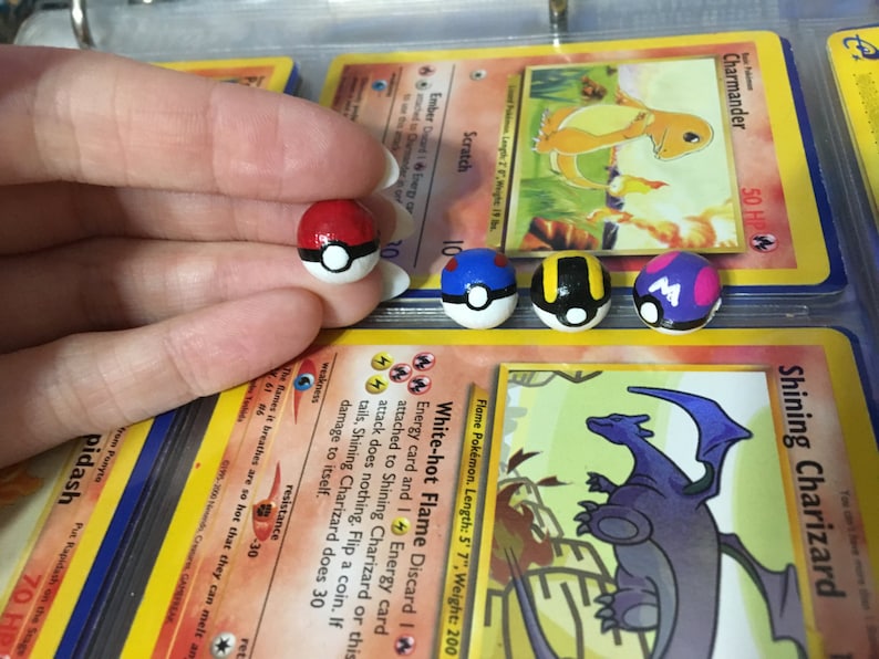 Op de afbeelding: Vier handgeschilderde Pok&eacute;mon-personage Pok&eacute; Balls. De Pok&eacute; Balls zijn rood, blauw, zwart en geel, en paars en roze. De Pok&eacute; Balls liggen op Pok&eacute;mon-verzamelkaarten.