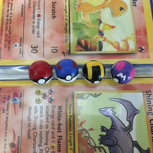 Op de afbeelding: Vier kleine, handgeschilderde, ronde Pokemon-figuren van keramiek. De figuren zijn geschilderd in de kleuren van de Pokemon-karakters: rood, blauw, zwart en paars. Elk figuur heeft een witte cirkel aan de voorkant met een zwart ontwerp.
