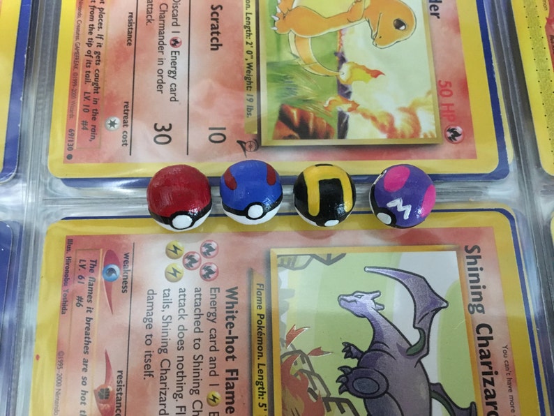 Op de afbeelding: Vier kleine, handgeschilderde, ronde Pokemon-figuren van keramiek. De figuren zijn geschilderd in de kleuren van de Pokemon-karakters: rood, blauw, zwart en paars. Elk figuur heeft een witte cirkel aan de voorkant met een zwart ontwerp.