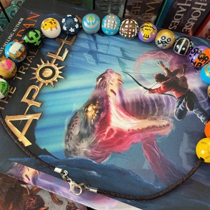 May include: A brown cord necklace with colorful wooden beads. Each bead is painted with a different symbol, including a tree, a star, a crescent moon, and a sun. The necklace is lying on top of a book titled "The Trials of Apollo: The Tower of Nero, Book Five".