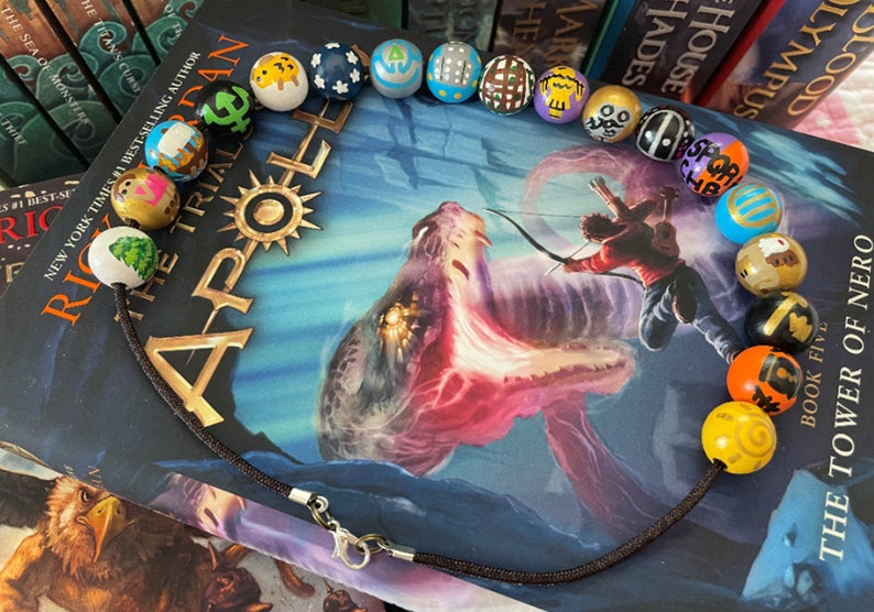 May include: A brown cord necklace with colorful wooden beads. Each bead is painted with a different symbol, including a tree, a star, a crescent moon, and a sun. The necklace is lying on top of a book titled "The Trials of Apollo: The Tower of Nero, Book Five".