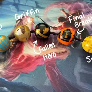 May include: A close-up of a beaded bracelet with five wooden beads. The beads are painted with different designs, including a harp, a griffin, a fallen hero, a final breath, and a sun.