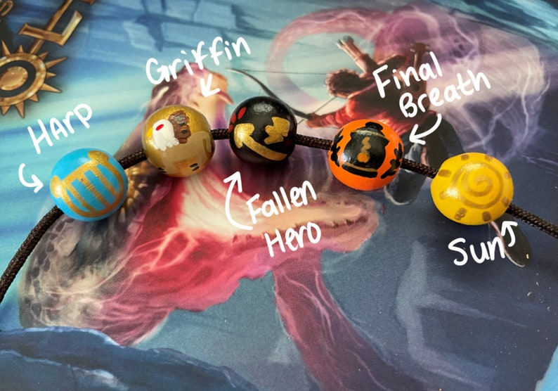 May include: A close-up of a beaded bracelet with five wooden beads. The beads are painted with different designs, including a harp, a griffin, a fallen hero, a final breath, and a sun.