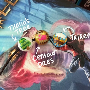 May include: A black cord with three wooden beads. The beads are painted with images of a tree, a centaur, and a ship. The beads are labeled "Thalia's Tree", "Centaur Dres", and "Trireme".
