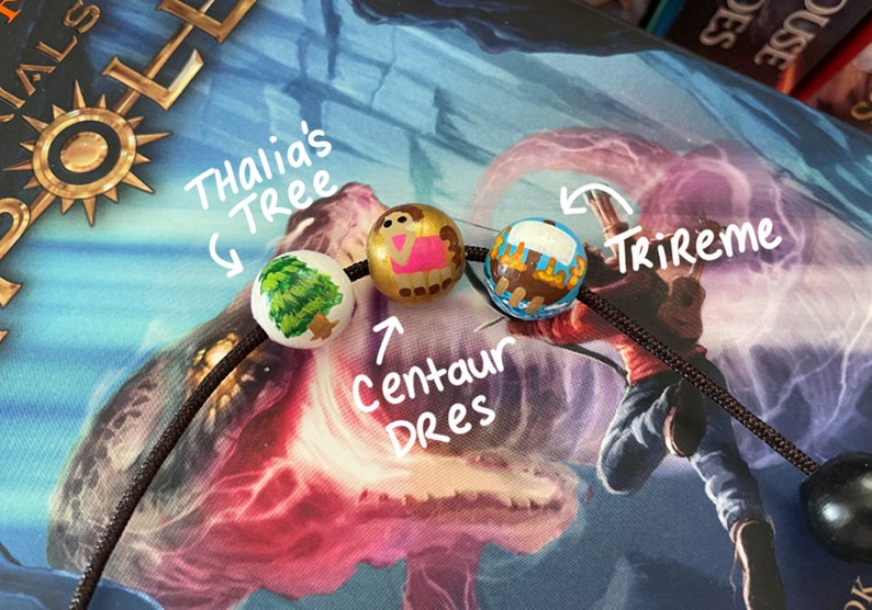 May include: A black cord with three wooden beads. The beads are painted with images of a tree, a centaur, and a ship. The beads are labeled "Thalia's Tree", "Centaur Dres", and "Trireme".