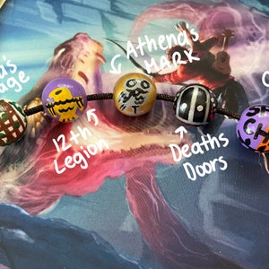 May include: A close-up of a beaded necklace with six colorful beads. The beads are decorated with various symbols and text, including "Hera's cage", "12th Legion", "Athena's Mark", "Deaths Doors", and "Camps Unite".