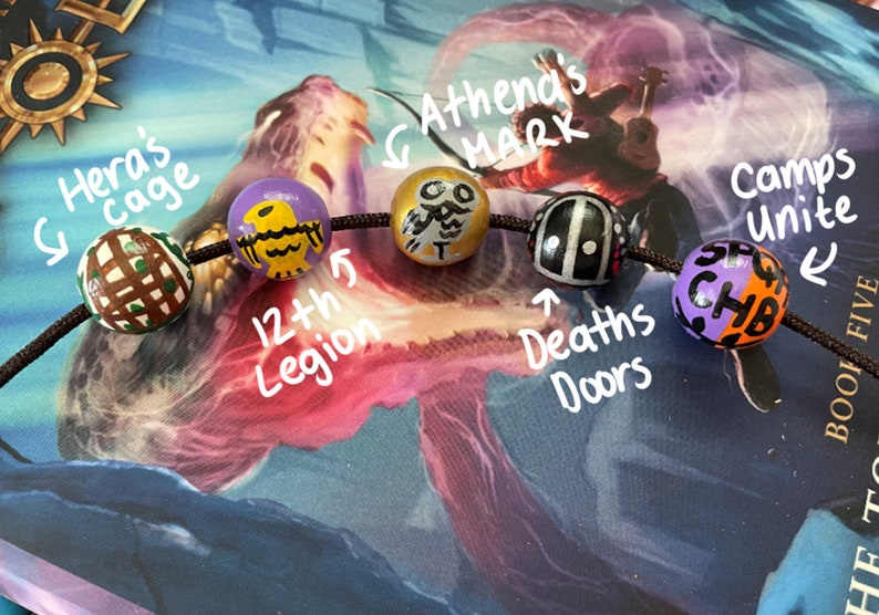 May include: A close-up of a beaded necklace with six colorful beads. The beads are decorated with various symbols and text, including "Hera's cage", "12th Legion", "Athena's Mark", "Deaths Doors", and "Camps Unite".