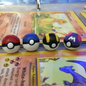 Op de afbeelding: Vier handgeschilderde Pokeball-versieringen, elk met een ander kleurenschema: rood en wit, blauw en wit, zwart en geel en paars en wit. De Pokeballs liggen op Pok&eacute;mon-verzamelkaarten.