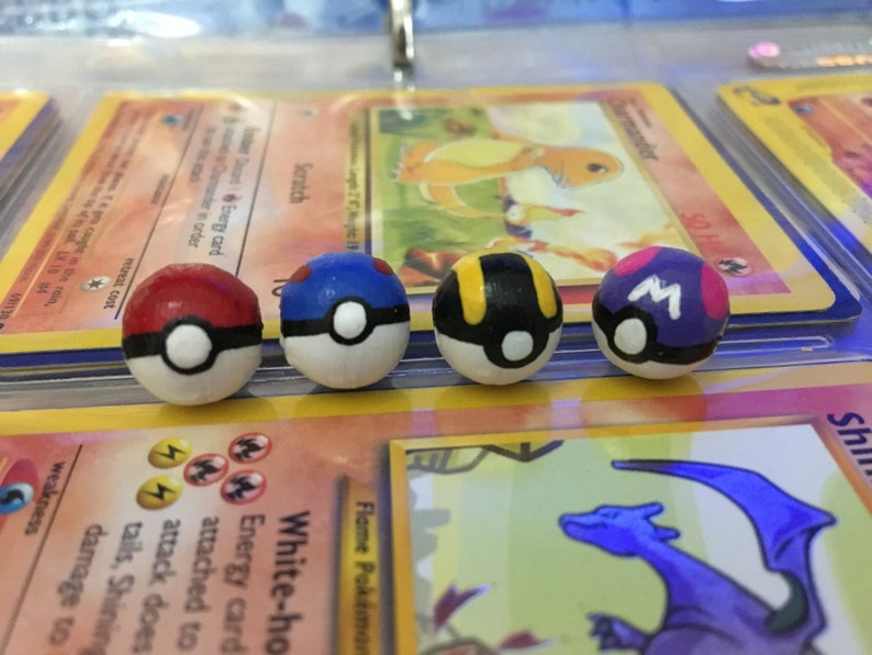 Op de afbeelding: Vier handgeschilderde Pokeball-versieringen, elk met een ander kleurenschema: rood en wit, blauw en wit, zwart en geel en paars en wit. De Pokeballs liggen op Pok&eacute;mon-verzamelkaarten.