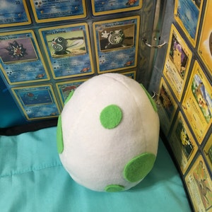 May include: A white plush egg with green polka dots. The egg is sitting on a blue surface in front of a binder filled with Pokemon trading cards.