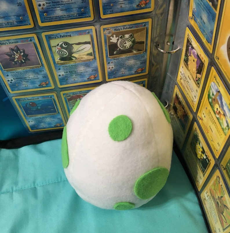 May include: A white plush egg with green polka dots. The egg is sitting on a blue surface in front of a binder filled with Pokemon trading cards.