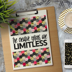 K&ouml;nnte beinhalten: Ein motivierendes Zitat, "the creative options are LIMITLESS", gedruckt auf einer wei&szlig;en Karte mit einem schwarzen und goldenen geometrischen Muster. Die Karte befindet sich auf einer Klemmbrett mit einem h&ouml;lzernen Hintergrund.