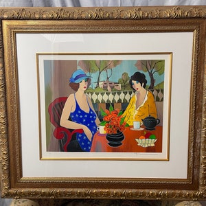 May include: A framed print of a painting depicting two women sitting at a table outdoors. The woman on the left is wearing a blue dress with white polka dots and a blue hat. The woman on the right is wearing a yellow dress. There are flowers and a teapot on the table.