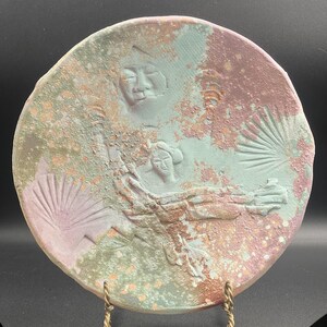 May include: A round, ceramic plate with a textured surface and a unique design. The plate features embossed figures and fan-like shapes in shades of green, pink, and brown. The plate is displayed on a gold-colored stand.