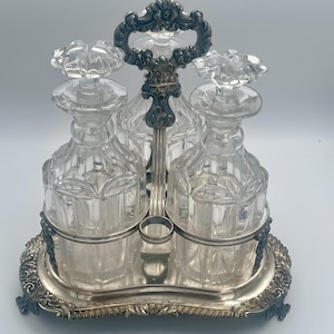 May include: A silver and crystal decanter set with three bottles and a decorative handle. The bottles have a cut glass design and sit on a silver tray with ornate feet.