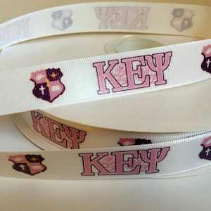 May include: White ribbon with pink lettering that reads "KEY" and a purple and gold crest with a cross.