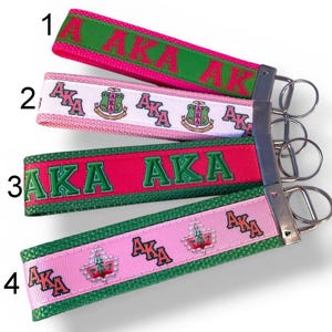 May include: Four keychains with fabric straps and metal key rings. The straps feature the letters "AKA" in various colors, including pink, green, and white. Some straps have the Greek letters and crests.
