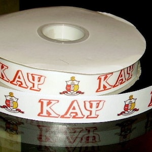 May include: A roll of white ribbon with red lettering that says "ΚΑΨ" and a red and gold crest. The ribbon is on a black surface.