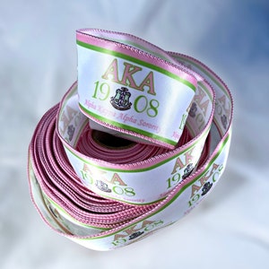 May include: A roll of white satin ribbon with pink and green trim. The ribbon has the text "AKA 1908 Alpha Kappa Alpha Sorority" printed on it.