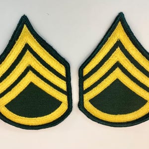 May include: Two Sergeant rank insignia patches. Each patch features three yellow chevrons on a dark green background, with a yellow border. The patches are symmetrical and designed for military uniforms.