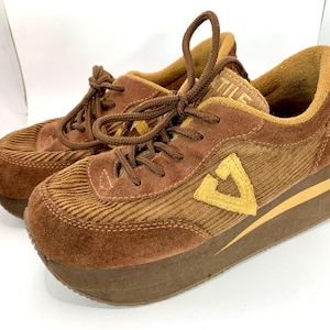 May include: A pair of brown platform sneakers featuring a combination of corduroy and suede textures. The shoes have brown laces, a thick brown platform sole with a thin tan stripe, and a yellow V-shaped logo on the side. The tongue displays the text TILE.