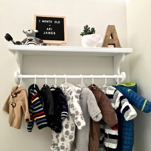 Puede incluir: Estante blanco con una barra de metal blanco para colgar ropa. El estante tiene un letrero que dice "1 month old Ari James". Hay 9 prendas de beb&eacute; colgadas en la barra, incluyendo un su&eacute;ter marr&oacute;n, un su&eacute;ter a rayas, un su&eacute;ter negro, un su&eacute;ter blanco, un su&eacute;ter gris, un su&eacute;ter marr&oacute;n, un su&eacute;ter a rayas, un su&eacute;ter blanco y un su&eacute;ter azul.