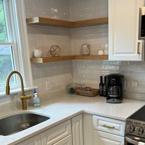 Puede incluir: Una cocina blanca con gabinetes blancos y una encimera blanca. Hay un grifo dorado sobre un fregadero de acero inoxidable. Hay dos estantes de madera en la pared sobre la encimera. Los estantes est&aacute;n llenos de varios art&iacute;culos, incluyendo una peque&ntilde;a figura de b&uacute;ho, un objeto geom&eacute;trico dorado y algunos frascos. Hay una cafetera en la encimera.