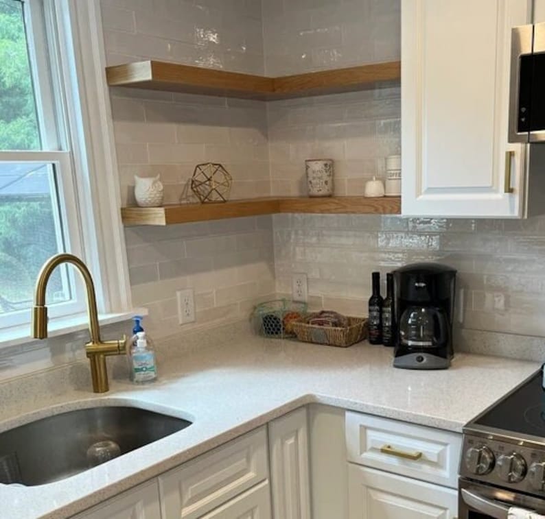 Puede incluir: Una cocina blanca con gabinetes blancos y una encimera blanca. Hay un grifo dorado sobre un fregadero de acero inoxidable. Hay dos estantes de madera en la pared sobre la encimera. Los estantes est&aacute;n llenos de varios art&iacute;culos, incluyendo una peque&ntilde;a figura de b&uacute;ho, un objeto geom&eacute;trico dorado y algunos frascos. Hay una cafetera en la encimera.