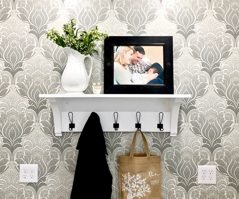 May include: A white wall-mounted shelf with four black metal hooks, featuring a white ceramic pitcher filled with green foliage and a black rectangular picture frame containing a photograph of a family, with a black coat and a tan tote bag hanging below.