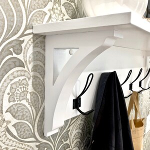 May include: A white wooden wall-mounted shelf featuring a flat top surface and two decorative support brackets. The unit includes a horizontal backboard equipped with four black metal hooks, one of which holds a dark fabric garment and a tan bag.