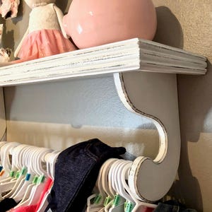 Puede incluir: Estante de madera blanca con acabado de pintura desgastada y soportes decorativos con volutas. El estante est&aacute; montado en una pared y tiene una barra de metal para colgar objetos.