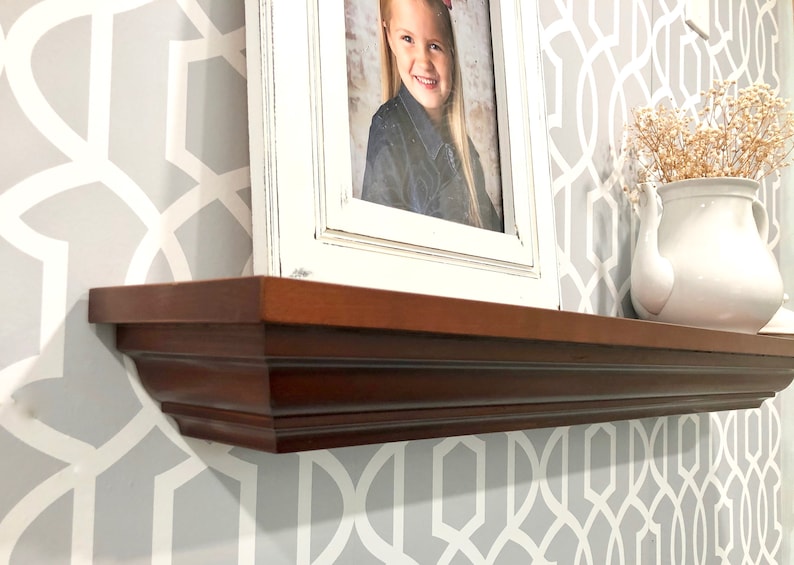 May include: A dark brown wooden shelf with a decorative molding detail. The shelf is mounted on a wall with a gray and white geometric wallpaper pattern. A white picture frame with a photo of a young girl is on the shelf. A white teapot with dried flowers is also on the shelf.