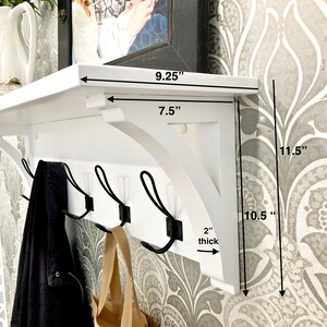 May include: A white wall-mounted shelf with four black metal hooks. The shelf top measures 9.25 inches wide, with a 7.5-inch deep bracket. The total height is 11.5 inches, the bracket height is 10.5 inches, and the shelf board is 2 inches thick.