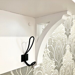 May include: A white wooden wall shelf featuring a curved support bracket and a black metal double hook attached to the front panel. The shelf is mounted against a wall with a grey and white patterned wallpaper design visible on the side.