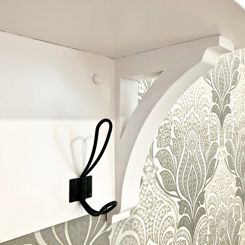 May include: A white wooden wall shelf featuring a curved support bracket and a black metal double hook attached to the front panel. The shelf is mounted against a wall with a grey and white patterned wallpaper design visible on the side.