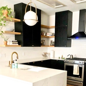Puede incluir: Una cocina moderna con gabinetes negros, salpicadero de azulejos blancos tipo metro y una estufa de acero inoxidable. La cocina cuenta con una isla grande con una encimera blanca y un grifo dorado. Hay estantes abiertos sobre los gabinetes con plantas y libros.