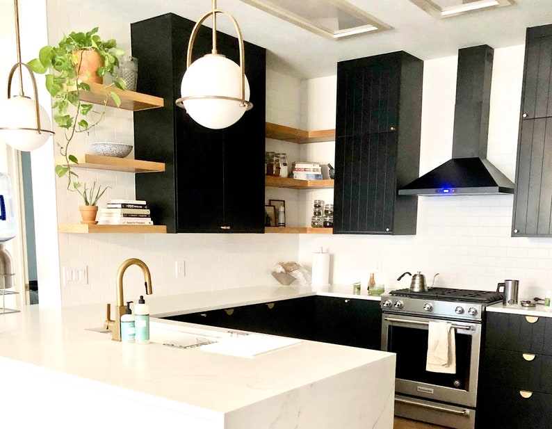 Puede incluir: Una cocina moderna con gabinetes negros, salpicadero de azulejos blancos tipo metro y una estufa de acero inoxidable. La cocina cuenta con una isla grande con una encimera blanca y un grifo dorado. Hay estantes abiertos sobre los gabinetes con plantas y libros.