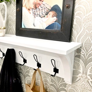 May include: A white wall-mounted shelf featuring three black metal hooks for hanging items. The shelf has a flat top surface and decorative brackets on the sides. A black wooden picture frame containing a photograph of a family is placed on top.